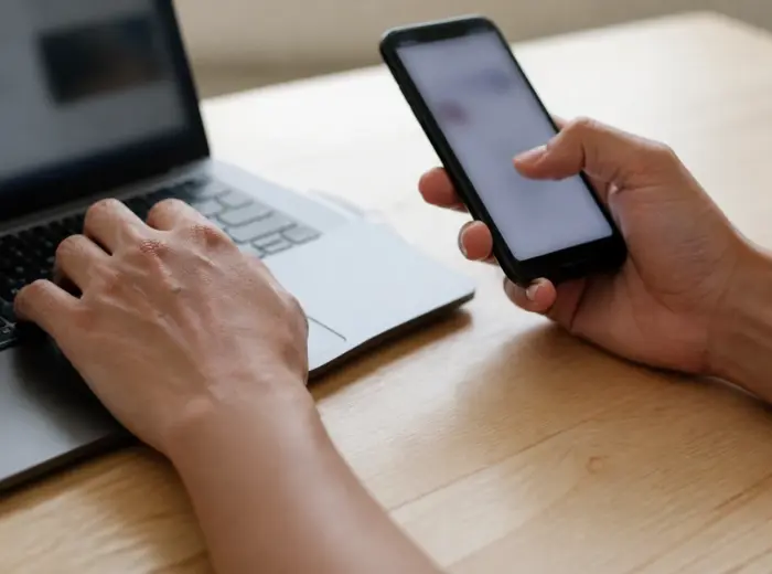 Gros plan de deux mains sur un bureau clair, la gauche sur un clavier, la droite tenant un téléphone