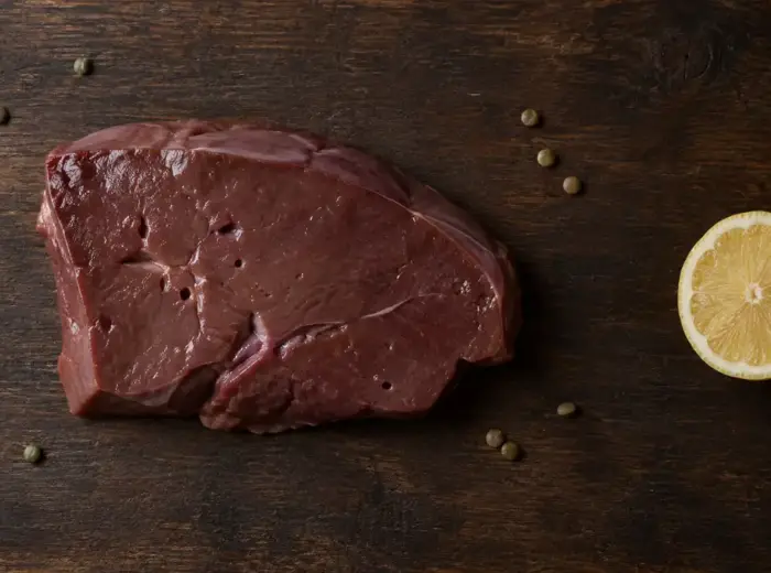 Tranche de foie de veau cru posée sur une planche en bois foncé, entourée de lentilles vertes et d'un demi-citron, vue de dessus